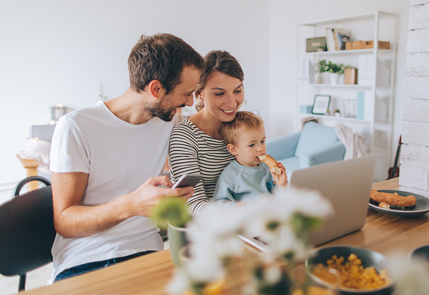 couple looking at a laptop while holding their infant and eating breakfast together